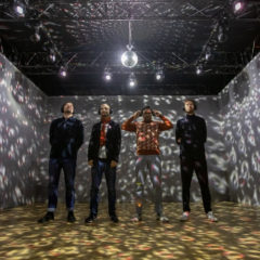 Four men in a room, with a disco ball on the ceiling and projections of headphones on the walls.