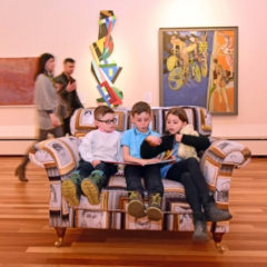 Three children sat on a sofa reading a book. Two adults are walking past the sofa.