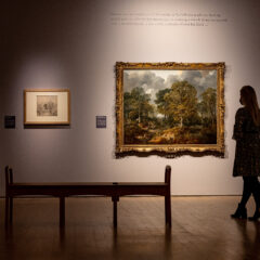 A back wall has 4 artworks displaying. two drawings on the left, followed by a large painting followed by a small drawing on the right. A woman stands admiring the painting in the foreground