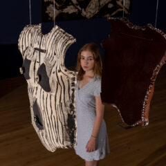 A visitor stands in the gallery beside a hanging artwork. The work looks like the breastplate of a suit of armor. The image is framed so that the figure appears between the two piece of the artwork.