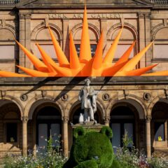 A view of the outside of the York Art Gallery with blooming flowers and pants in the foreground and the façade of the gallery in the background, with Steve Messam's spikey orange installation piece visible over the entrance to the building.